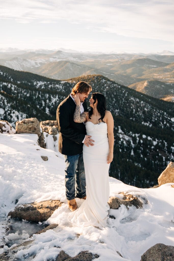 groom hugging bride from behind as he touches his face as the sun sets on top of a mountain during their boulder elopement