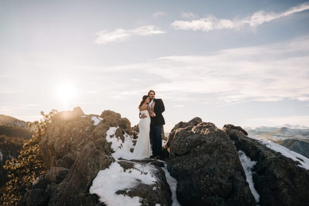 A couple in wedding attire stands on rocky, snow-dusted terrain with mountains in the background and the sun setting, creating a warm, romantic atmosphere.