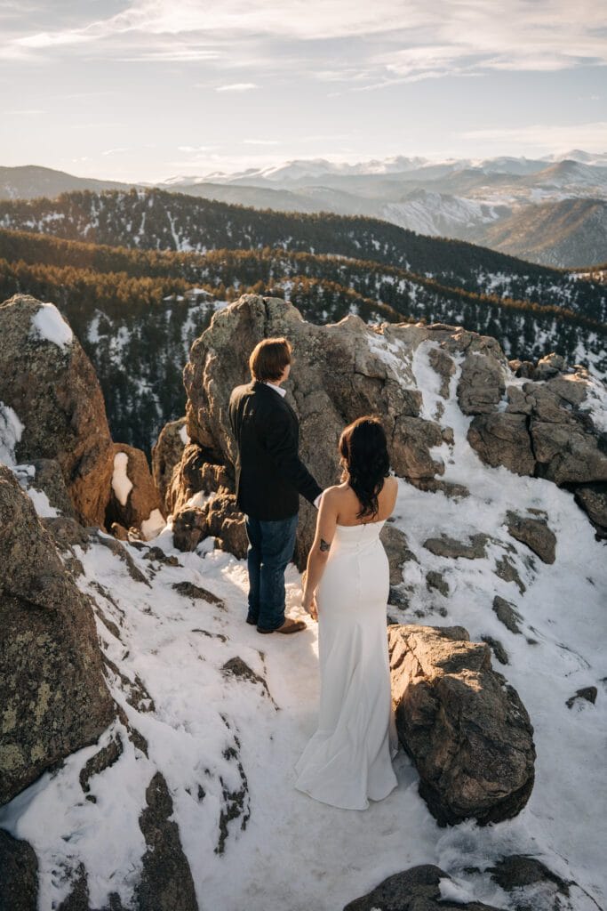 A couple, with the woman in a white dress and the man in a dark jacket, stand on a snowy rocky mountain ledge, overlooking a scenic view of forested hills and distant snow-capped mountains under a cloudy sky.