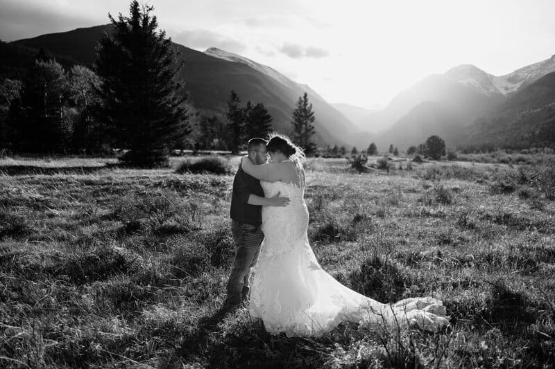 A couple embraces in a grassy mountain meadow at sunset, with the bride in a long white gown and the groom in casual attire. Tall trees and peaks surround them under a dramatic sky. The scene is in black and white.