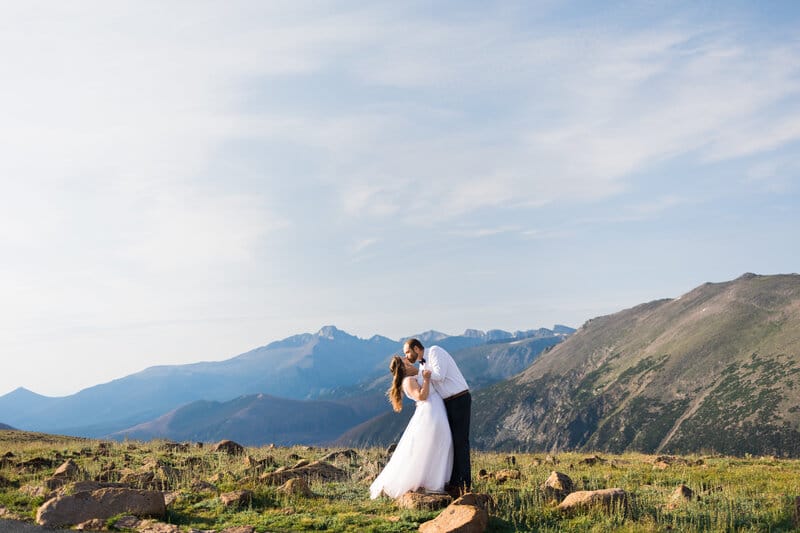 A bride and groom kiss on a grassy mountaintop with rocky terrain. Majestic mountains and a blue sky with wispy clouds form the backdrop.