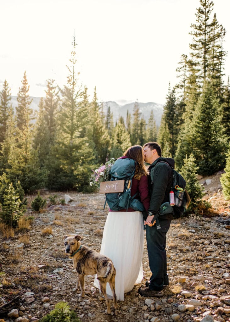 A couple dressed for hiking, with backpacks and wedding attire, kiss on a forest trail surrounded by pine trees. A tan dog stands in the foreground, and mountains are visible in the background.