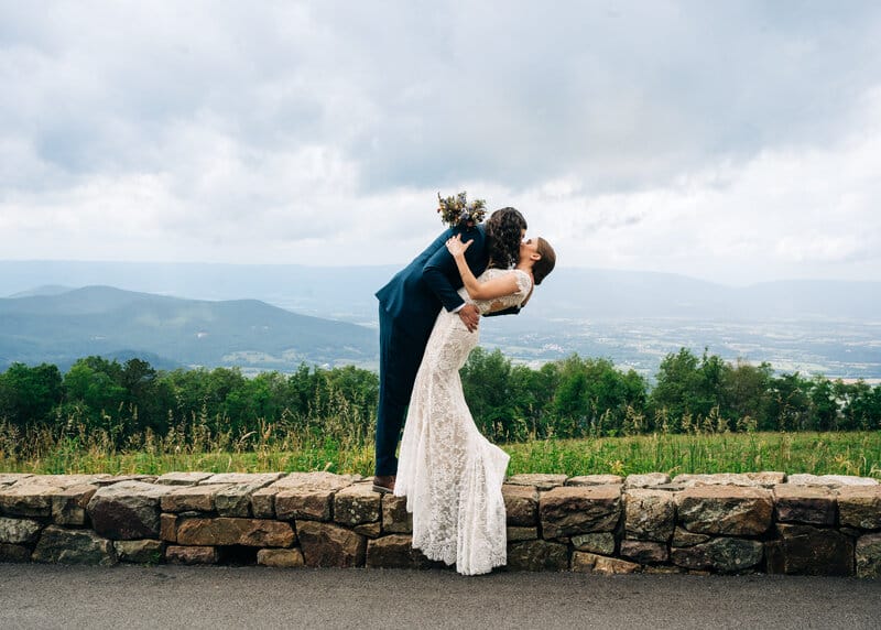 A groom in a blue suit dips and kisses a bride in a white lace dress on a stone wall, with mountains, greenery, and cloudy sky in the background.