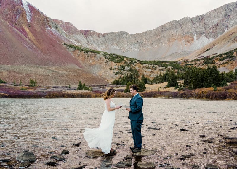 A bride and groom stand on rocks in a shallow lake, holding hands, surrounded by colorful mountains and pine trees under an overcast sky. The brides white dress flows in the breeze.