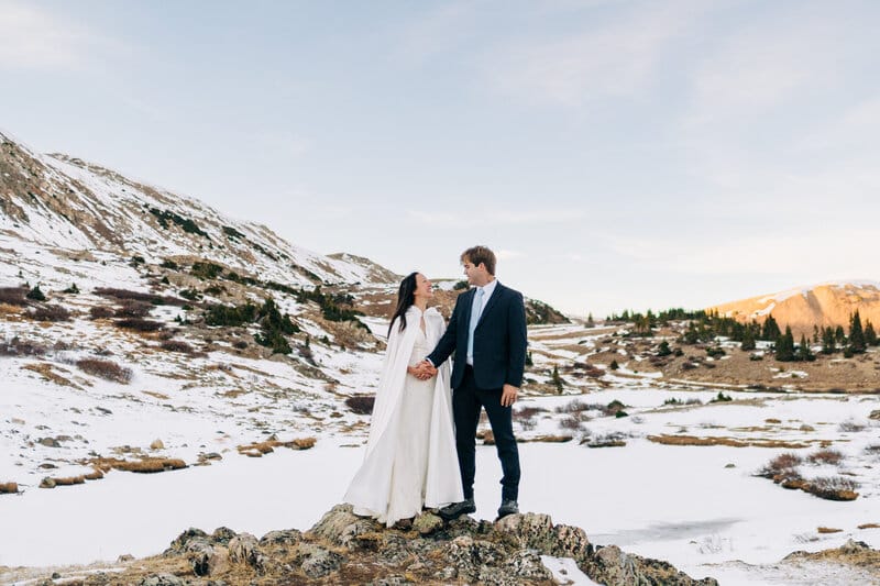 A couple dressed in formal attire stands on rocky ground in a snowy mountain landscape, holding hands and looking at each other under a clear sky.