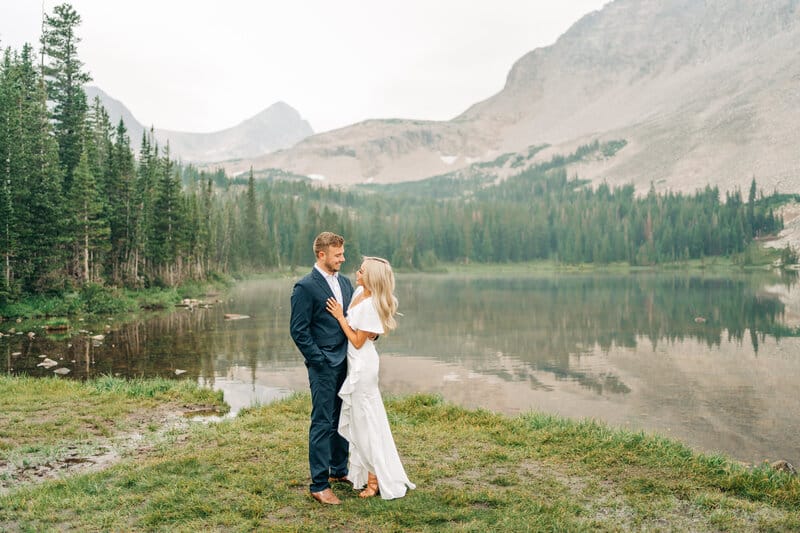 A couple stands by a calm mountain lake, surrounded by pine trees and misty mountains. The man wears a dark suit, and the woman wears a white dress. They smile at each other, embracing on the grassy shore.