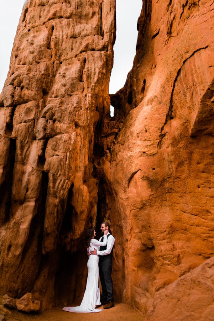 A bride and groom embrace at the base of towering, textured red rock formations, surrounded by dramatic cliffs. The bride wears a white dress, and the groom is dressed in a vest and tie.