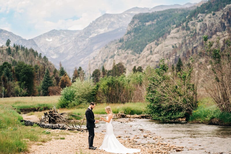 A bride and groom stand facing each other by a river, surrounded by lush greenery and mountains in the background under a partly cloudy sky.