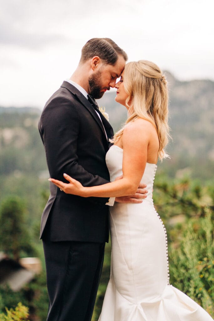 A bride and groom stand outdoors, embracing and touching foreheads, smiling softly. The bride wears a strapless white dress; the groom is in a black suit. Greenery and blurred mountains are in the background.