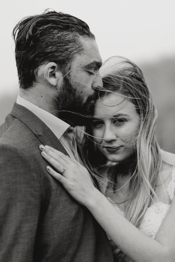 A black-and-white photo of a couple embracing; the man in a suit gently rests his forehead on the womans head, while she looks at the camera, her hand placed on his chest, both appearing intimate and serene.