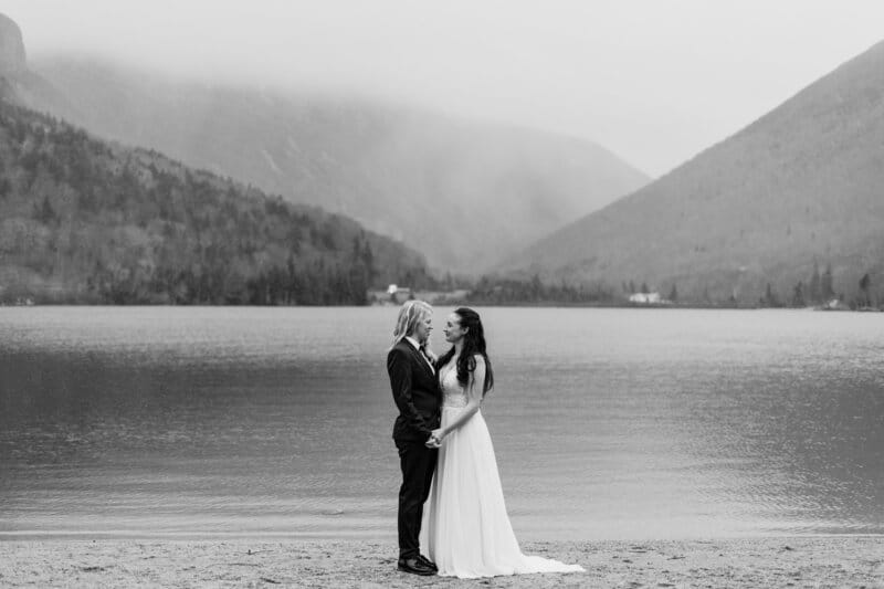 A bride and groom stand on a sandy shore holding hands, gazing at each other, with a calm lake and misty, forested mountains in the background. The image is in black and white.
