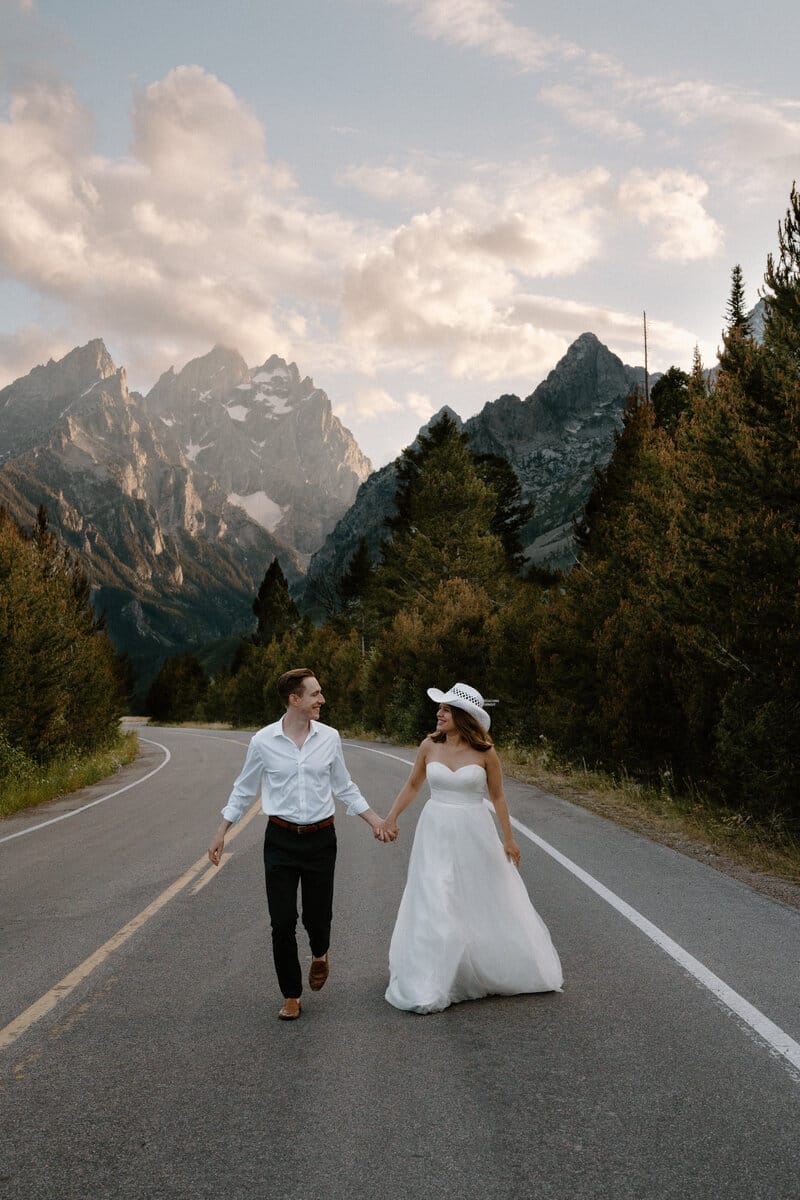 A bride and groom hold hands while walking on a winding mountain road, surrounded by pine trees and dramatic peaks under a cloudy sky at sunset. The bride wears a white dress and hat; the groom is in a white shirt and dark pants.