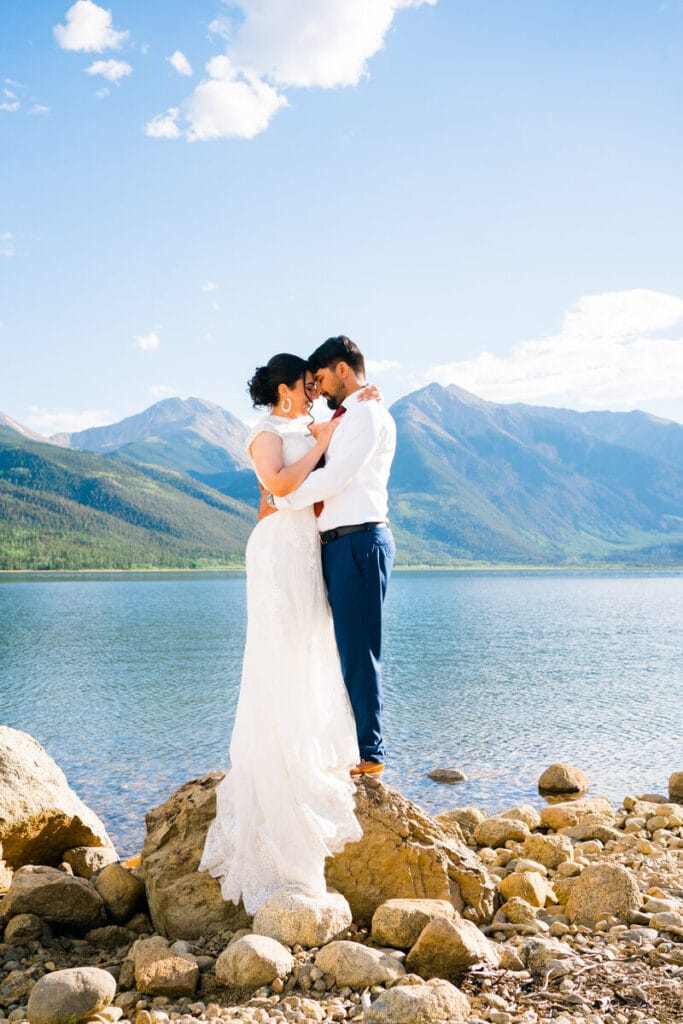 A couple dressed in wedding attire embrace on rocks beside a serene lake, with mountains and a blue sky with scattered clouds in the background.
