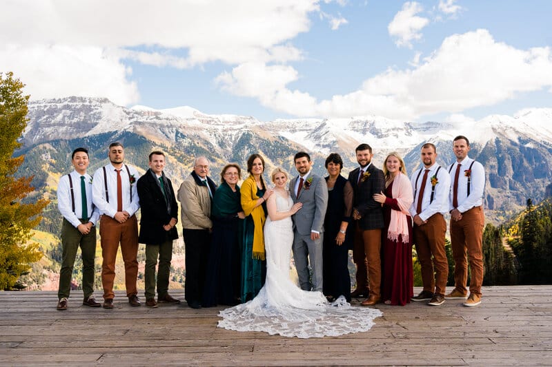 A wedding party poses outdoors on a wooden deck with snowy mountains in the background. The bride and groom stand at the center, surrounded by family and friends dressed in formal and semi-formal attire.