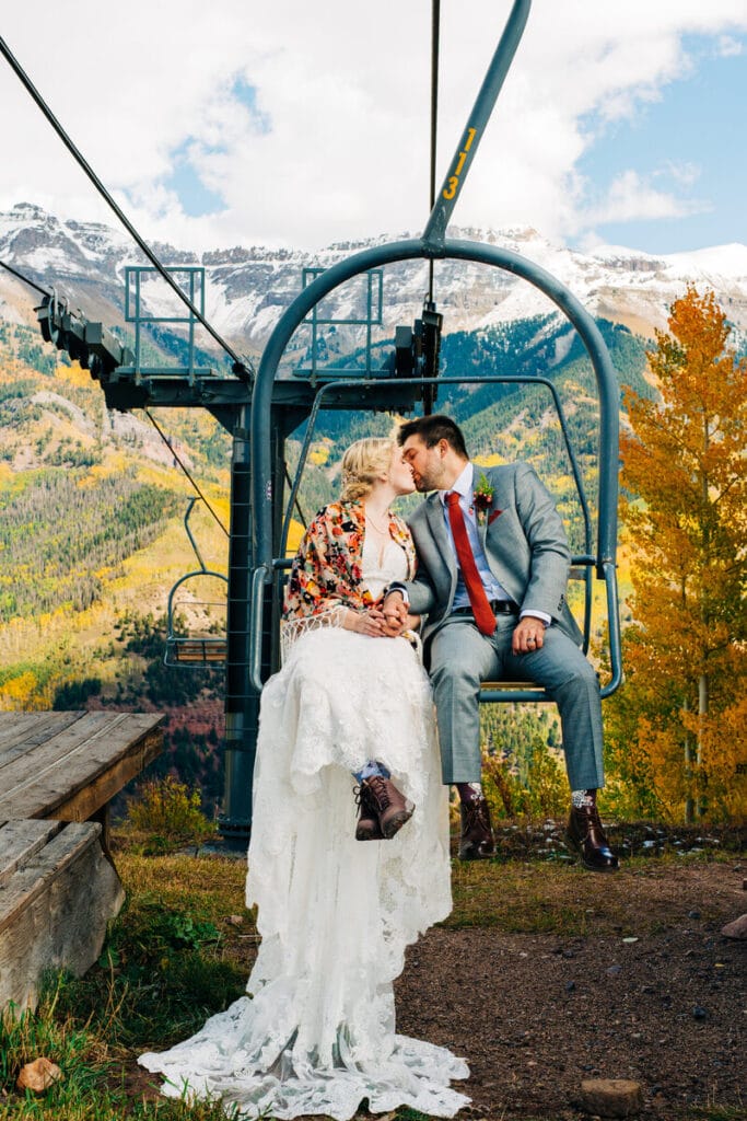 A bride and groom sit on a ski lift, sharing a tender moment. The bride wears a white dress and floral shawl, while the groom is in a grey suit and red tie. Mountains and autumn trees fill the scenic background.
