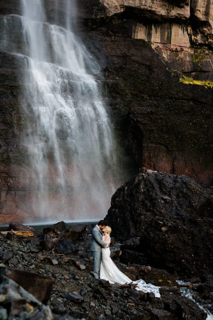 A bride and groom embrace at the base of a towering waterfall, surrounded by rugged rocks and mist, creating a dramatic and romantic outdoor wedding scene.