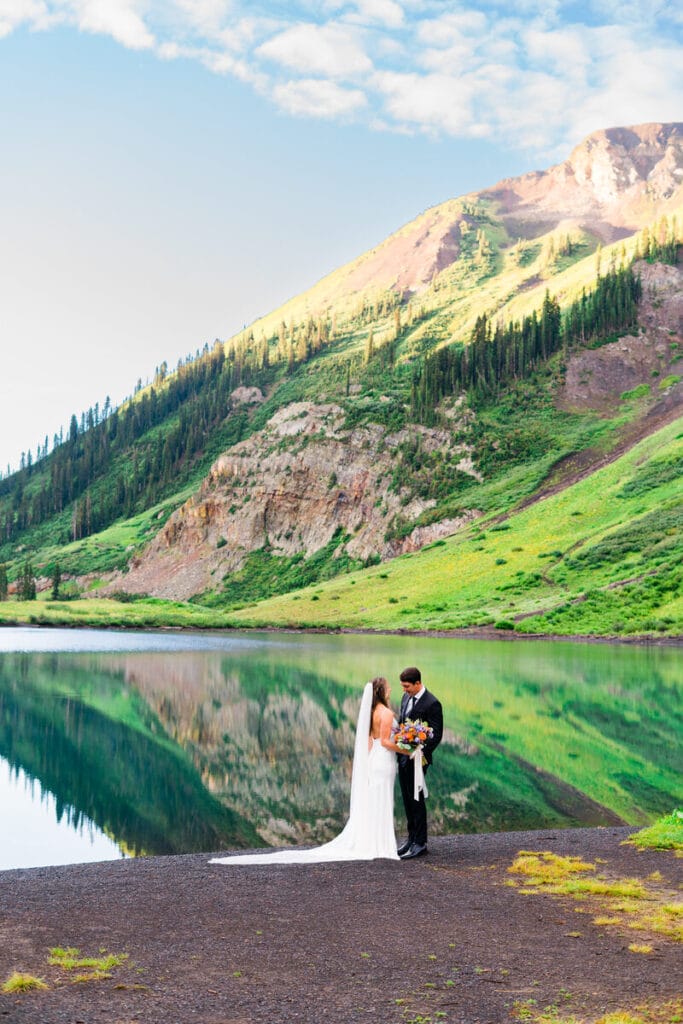 A bride and groom stand together by a calm mountain lake, with green hills and a sunlit peak reflected in the water under a partly cloudy sky. The bride wears a white dress and veil, and the groom holds a bouquet.
