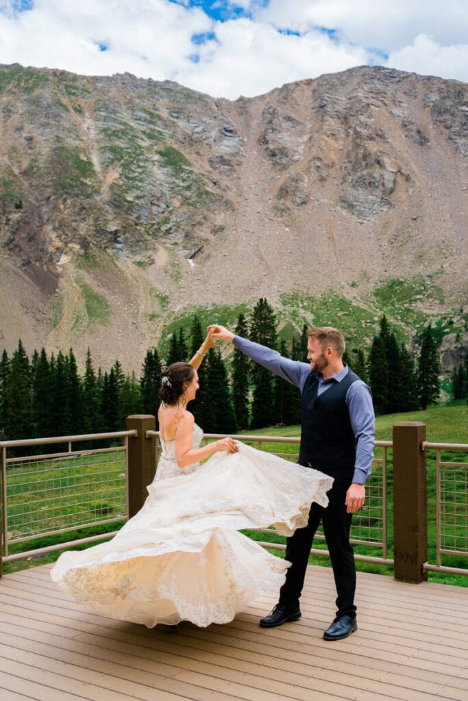 A bride and groom dance on a wooden deck with green pine trees and a rocky mountain in the background. The bride’s dress twirls elegantly as she spins, and both appear joyful against the scenic landscape.