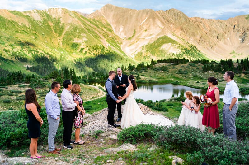 A bride and groom exchange vows outdoors on a mountain, surrounded by friends and family. A small lake and green hills are in the background under a blue sky. The wedding party stands in a semicircle, watching the couple.
