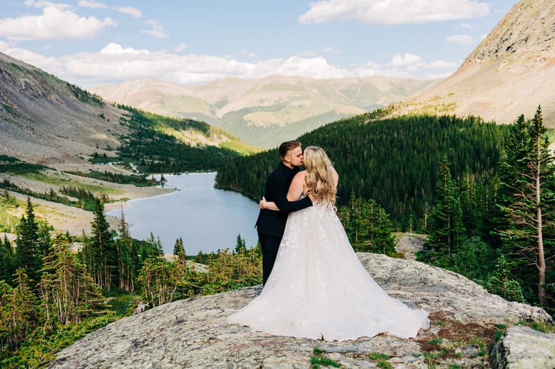 A couple in wedding attire embraces on a rocky cliff overlooking a scenic mountain landscape with a forest and a lake under a partly cloudy sky.