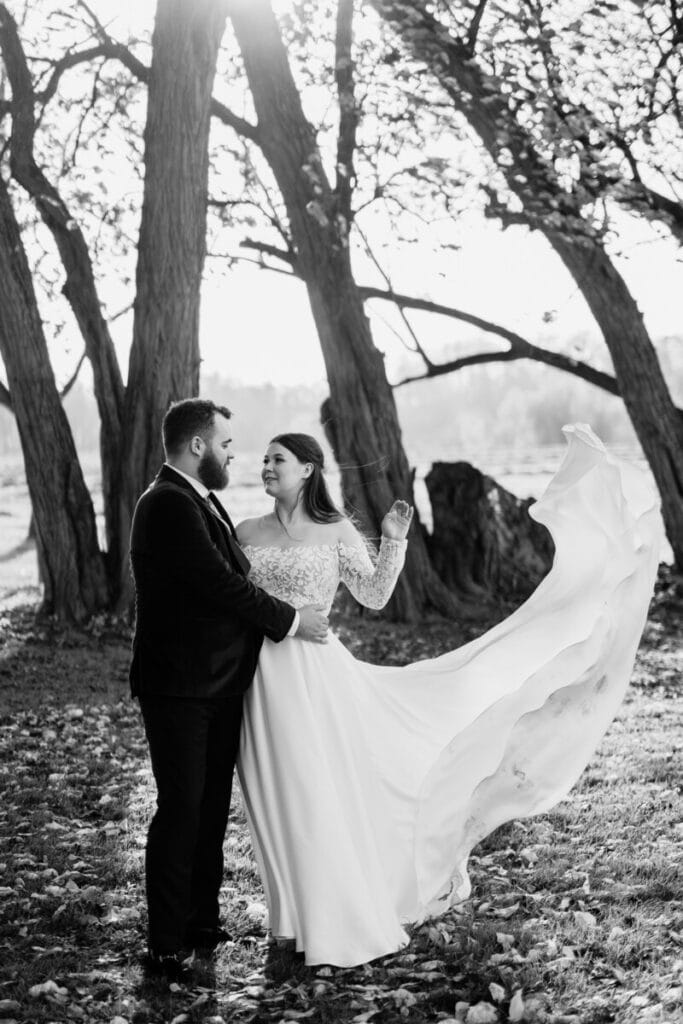 A bride and groom stand together outdoors under tall trees. The groom holds the bride, whose long dress flows dramatically in the wind. Sunlight filters through the branches, creating a romantic, serene scene. Black and white photo.