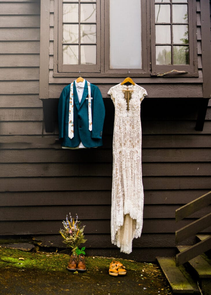 A lace wedding dress and a teal suit with a floral tie hang side by side under a window on a dark wooden wall. Below them are brown shoes, tan sandals, and a bouquet of flowers.