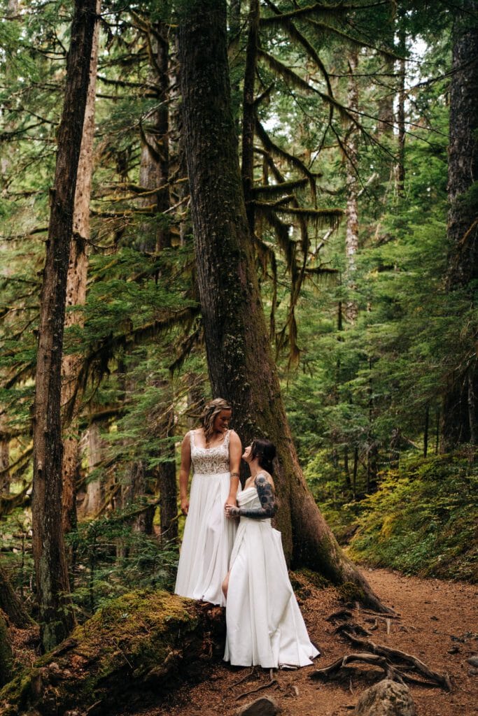 Two women in white dresses stand together on a forest path surrounded by tall trees and lush greenery, sharing a joyful moment in a natural, outdoor setting.