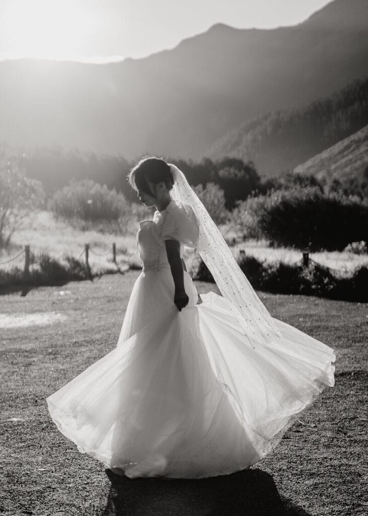 A bride in a flowing wedding dress and veil twirls outdoors at sunset, with mountains and trees silhouetted in the background. The scene is in black and white, creating a dreamy and romantic atmosphere.