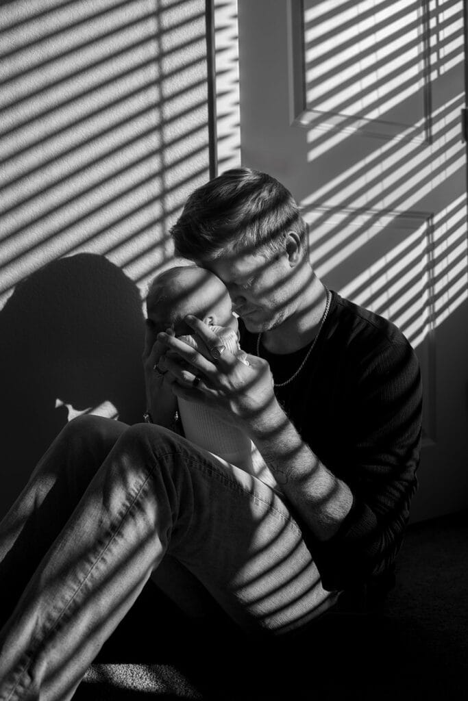 A man sits on the floor holding a baby, both illuminated by sunlight streaming through window blinds. Striped shadows dance across their faces and bodies, capturing a moment any newborn photographer would cherish.