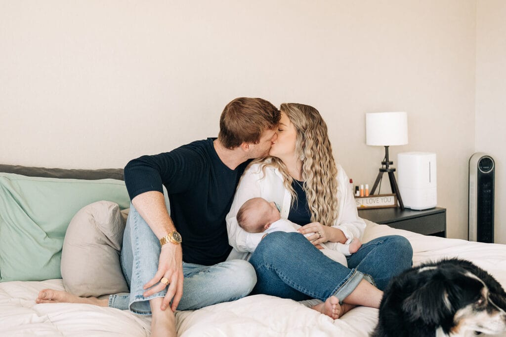 A couple sits on a bed, kissing, while the woman holds a baby in her arms. Captured by a newborn photographer, a black dog rests at the edge of the bed. A nightstand with a lamp and an I love you sign complete the cozy scene.