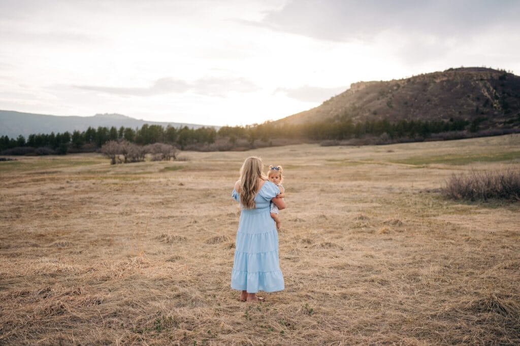 A woman in a light blue dress stands barefoot in a dry, grassy field, holding a young child. They face away towards distant hills and a cloudy sunset sky of Dawson Butte—an intimate moment perfect for Family Photography. Sparse trees line the horizon.