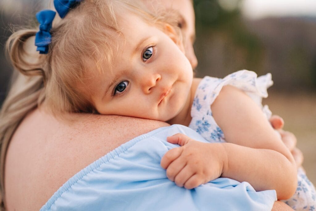 A toddler with blue eyes and a blue bow in her hair rests her head on an adults shoulder, gazing softly at the camera. She wears a light blue dress with floral patterns.