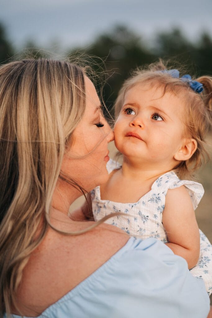 A woman with blonde hair holds a young child in a blue and white dress and bow. They are outdoors, surrounded by blurry trees, sharing a tender moment—a perfect example of heartfelt family photography.
