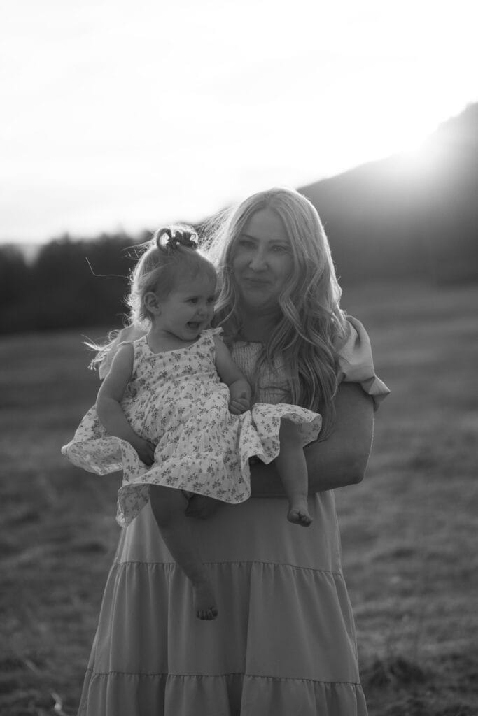 A woman with long hair holds a young girl in a floral dress outdoors in a grassy field at sunset. Both look slightly away from the camera, capturing a timeless moment of family photography in black and white.