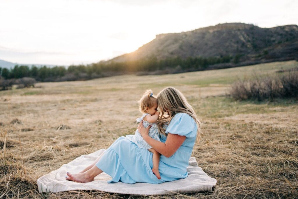 A woman in a light blue dress sits on a blanket in a grassy field at Dawson Butte, holding a young child. The sun sets behind distant hills, creating a warm and peaceful Mommy and Me Photoshoot scene.