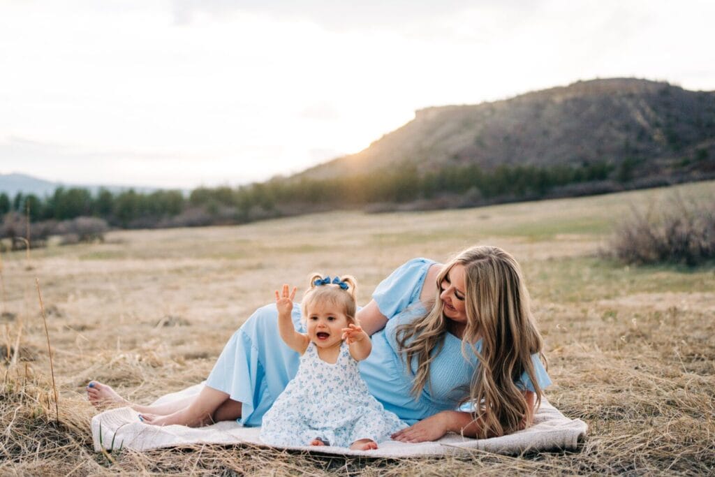 A denver family photographer snaps a shot of a woman in a light blue dress lies on a blanket in a grassy field, smiling at a toddler girl in a matching blue dress with arms raised. Hills and trees are visible at sunset—capturing a joyful Family Photography moment.