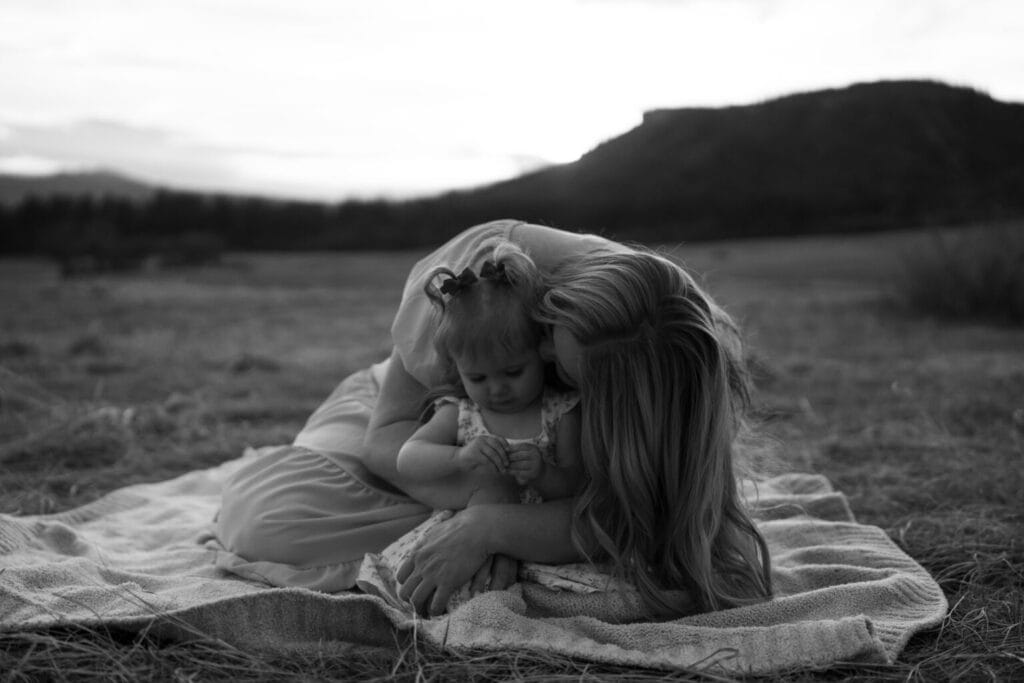 A woman hugs and kisses a young child on the head while sitting on a blanket in an open field, with hills and trees in the background. Captured in black and white by a Denver family photographer.