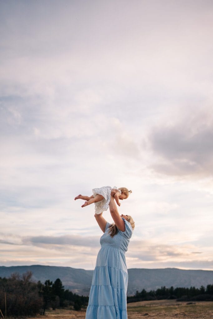 A woman in a light blue dress lifts a smiling young child above her head outdoors during a Mommy and Me Photoshoot, with the scenic mountains and trees of Castle Rock in the background under a cloudy sky.