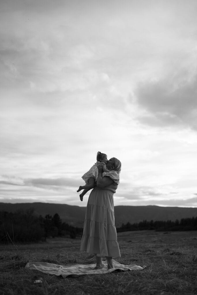 A woman in a long dress stands barefoot on a blanket in a field near Castle Rock, Colorado, lifting her child in a loving Mommy and Me Photoshoot as mountains rise beneath the cloudy sky.