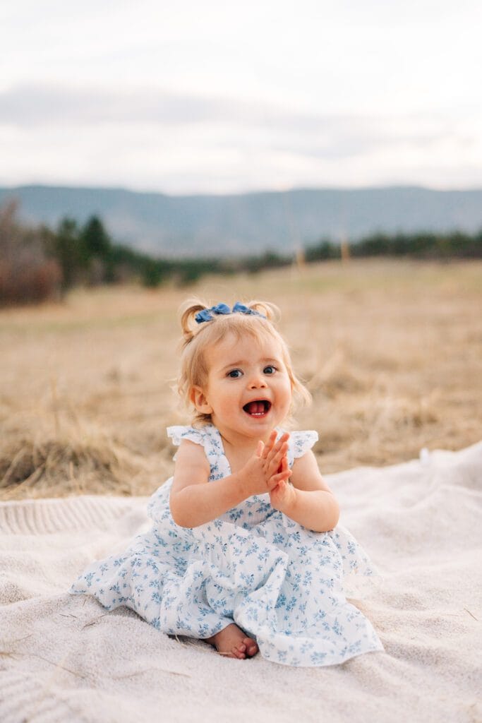 A young toddler with blonde hair in pigtails, wearing a white dress with blue flowers, sits on a blanket outdoors in a grassy field near Dawson Butte, smiling and clapping hands. Mountains and trees are visible in the background.