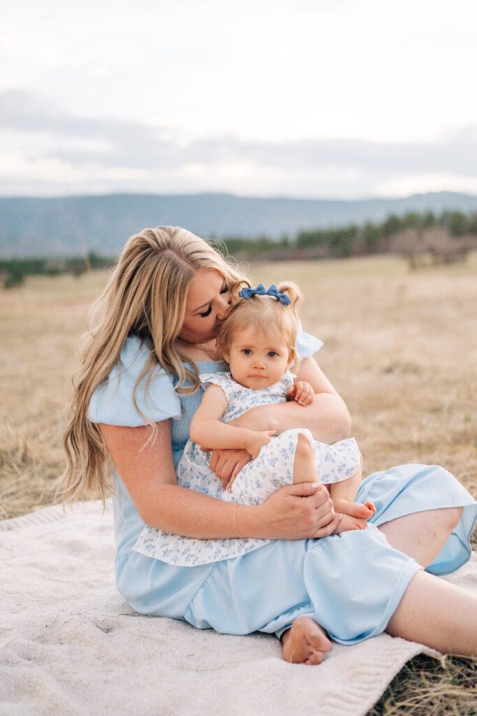 A woman in a light blue dress sits on a blanket in a field at Dawson Butte, hugging and kissing a toddler in matching blue and white for their Mommy and Me Photoshoot, with Castle Rock, Colorado’s mountains and trees in the background.