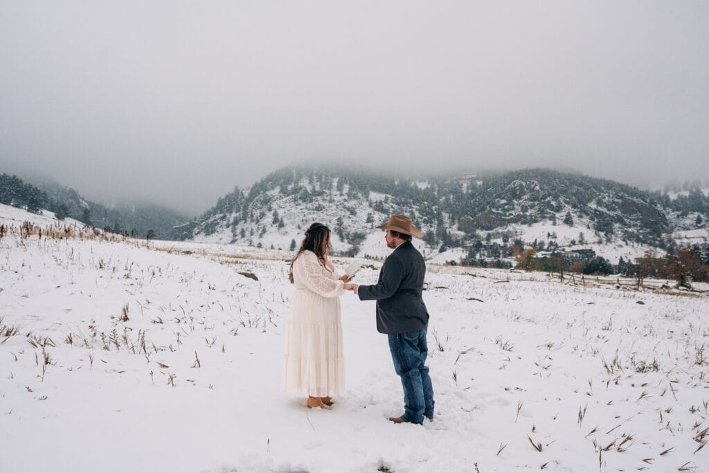 A couple stands facing each other in a snowy field with mountains in the background. The woman wears a long white dress, and the man wears a cowboy hat, jeans, and a jacket. The scene is overcast and peaceful.