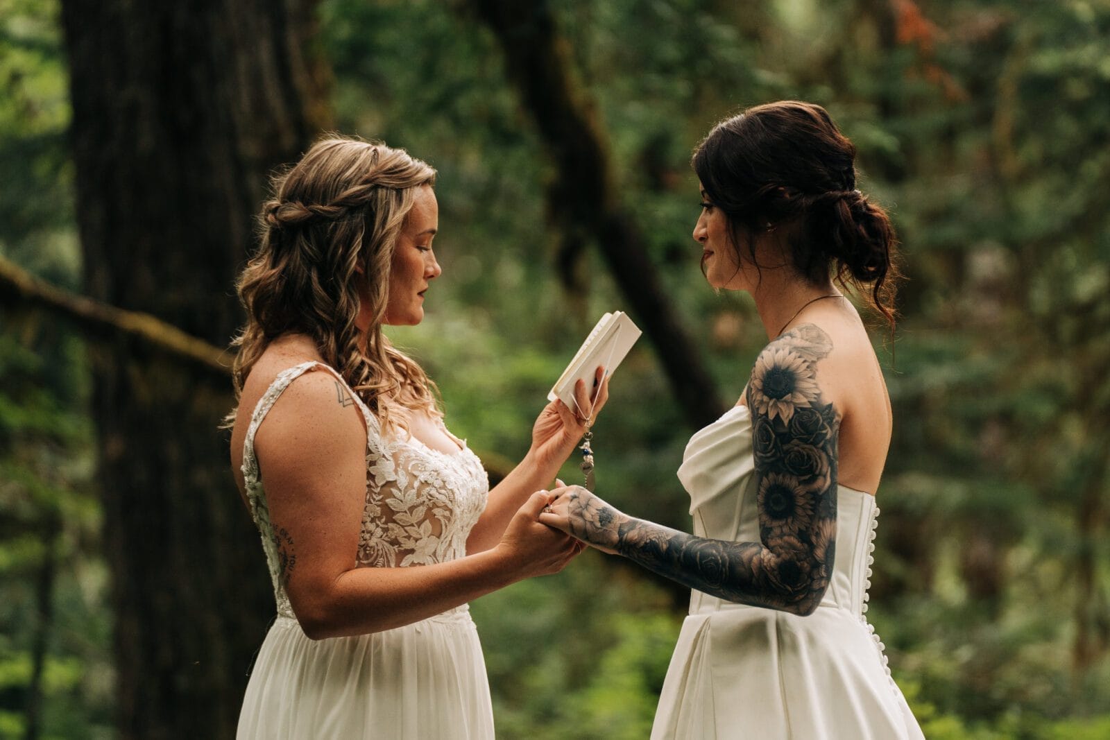 Two brides in white dresses hold hands in a forest. One bride with braided hair reads from a small book, while the other, with dark hair and floral tattoos, listens. Lush green trees surround them.