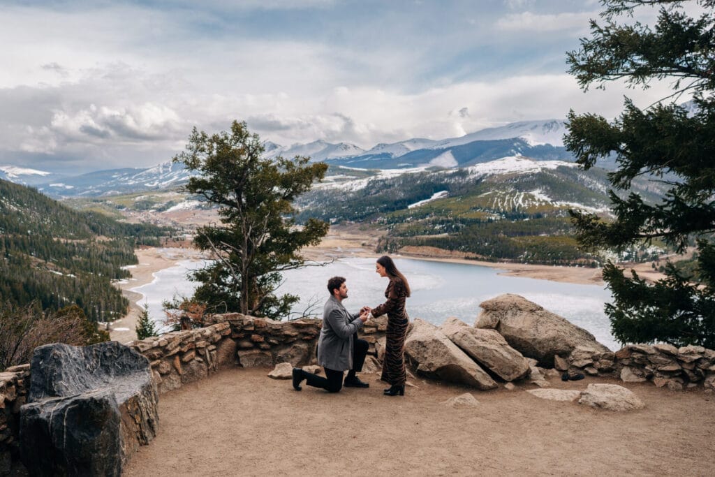 Eric down on his knee proposing to his girlfriend at Sapphire Point Overlook with the ten mile range in the background along with Dillon Reservoir in Breckenridge, Colorado
